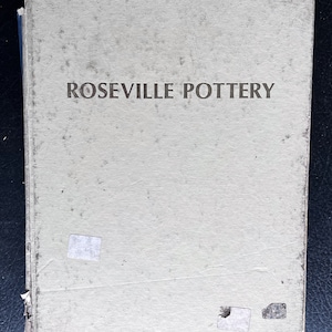 May include: A vintage book titled "Roseville Pottery" with a light green cover. The cover shows signs of wear, with visible stains and small sticker remnants. The title is printed in a dark brown font.