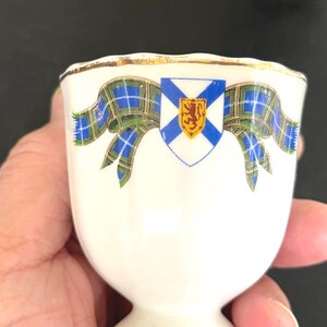 May include: A white ceramic egg cup featuring a gold-trimmed scalloped rim. The front displays a blue and green tartan ribbon graphic centered with a white shield containing a blue saltire cross and a small yellow shield with a red lion.