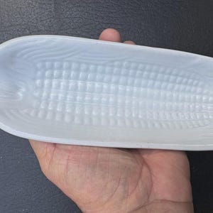May include: A white, corn-shaped serving dish. The dish is held in a hand and has a textured surface that mimics the appearance of corn kernels. The dish is made of a glossy material.
