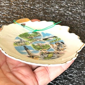 May include: A white ceramic souvenir dish with a scalloped gold-trimmed edge. The center features a collage of illustrations including Mt. Greylock, Elk Memorial, and Hail to the Sunrise, with text labels for each location and a green hanging string.