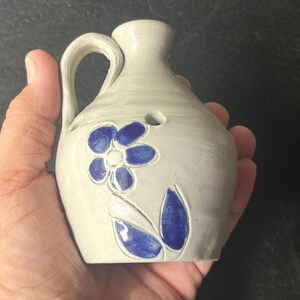 May include: A small, off-white ceramic pitcher with a handle and a narrow spout. The pitcher is decorated with a blue flower and leaf design. The pitcher is held in a person's hand.