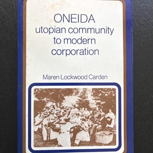 May include: A paperback book titled Oneida Utopian Community to Modern Corporation by Maren Lockwood Carden. The cover features a white background with a blue border and a brown frame containing a sepia-toned historical photograph of a group.