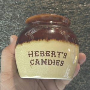 May include: A small, vintage ceramic jar with a brown and tan glaze. The jar features the text "HEBERT'S CANDIES" in brown lettering. The jar is held in a hand.