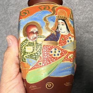 May include: A rectangular ceramic vase with a dark brown base and a hand-painted scene featuring two figures in traditional attire. The artwork includes vibrant orange, blue, green, and gold accents, with a dark rim at the top opening of the vessel.