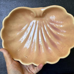 May include: A glossy, peach-colored ceramic dish shaped like a large scallop shell. The bowl features a scalloped edge and raised, radiating ridges that extend from a central hinge detail, with subtle purple-toned shading between the ridges.