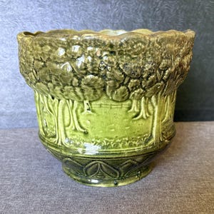 May include: A glazed ceramic planter featuring a relief design of a forest landscape with trees and foliage in shades of olive and lime green. The vessel has a scalloped top rim and a decorative base with a repeating geometric pattern.