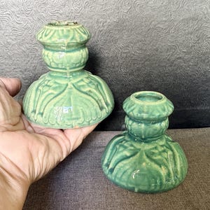 May include: Two sage green ceramic candle holders with a glossy finish and a molded, ribbed design. Each piece features a wide, rounded base that tapers upward into a smaller, circular top opening, showcasing a consistent, textured pattern.