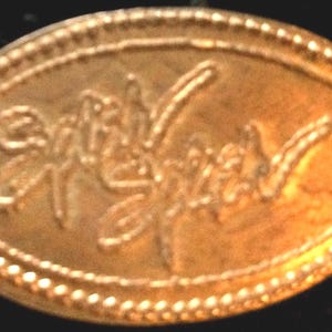 May include: A gold-colored oval-shaped coin or token with an embossed floral design and the word "Symphony" in script. The edge is decorated with a dotted border. The coin has a slightly worn appearance.