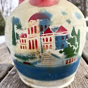 May include: A cream-colored ceramic jug featuring a painted scene of buildings with red roofs and blue walls, surrounded by green trees and a blue body of water. The design includes a prominent red dome and architectural details in red and blue.