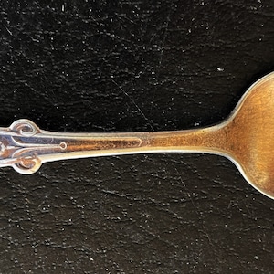 May include: A vintage silver spoon with an ornate handle and a rounded bowl. The handle features decorative floral and scrollwork details. The spoon is made of a shiny, reflective metal, and is likely used for serving or eating.