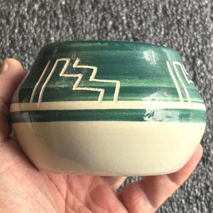 May include: A small, rounded ceramic pot featuring a two-tone design. The upper portion is glazed in a deep forest green with a hand-etched white geometric step pattern, while the lower section remains a smooth, unglazed off-white cream color.