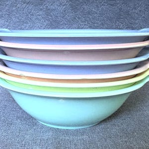 May include: A stack of six round bowls with integrated side handles, arranged in a vertical column. The bowls feature a smooth finish and are colored in a sequence of light blue, soft pink, periwinkle blue, beige, pale yellow, and light green.
