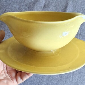 May include: A solid yellow gravy boat with a smooth, matte finish, featuring a wide, curved pouring spout and a matching integrated circular base plate. The vessel has a deep, rounded bowl shape and a clean, uniform color throughout the entire piece.