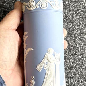 May include: A light blue ceramic vase with white relief detailing. The vase features a classical design with a woman playing a harp and floral patterns. The vase is cylindrical and has a decorative rim.