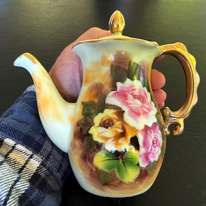 May include: A white porcelain teapot with a gold handle and lid. The teapot is decorated with a floral pattern of pink, yellow, and green roses.