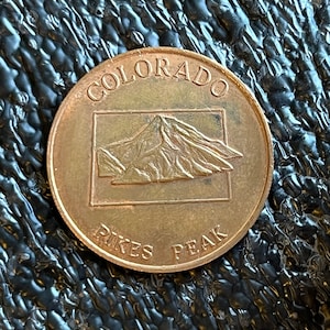 May include: A bronze-colored coin with the words "Colorado" and "Pikes Peak" inscribed around the edge. The center of the coin features a raised image of Pikes Peak.