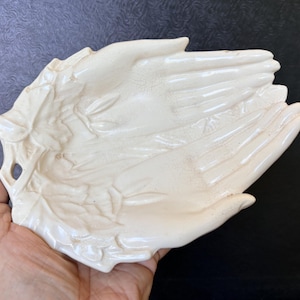May include: A cream-colored ceramic trinket dish shaped like two cupped hands with fingers extended. The surface features a glossy finish with subtle crackle detailing and embossed leaf-like patterns along the outer edges of the wrist area.