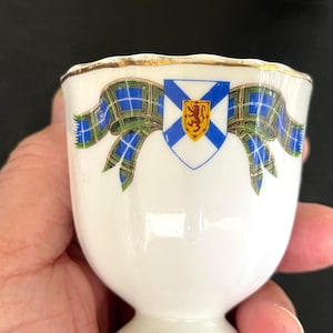 May include: White ceramic egg cup with a gold rim and a decorative design. The design features a blue and green plaid ribbon and a shield with a lion and a blue and yellow background. The egg cup is held in a hand.