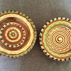 May include: Two round ceramic bowls featuring hand-painted designs in shades of terracotta, cream, and green. The left bowl displays concentric circles and dots, while the right bowl features a central spiral pattern, both with scalloped edges.