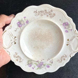 May include: A cream-colored ceramic serving plate with a scalloped edge and two integrated handles. The plate features a central crackle glaze finish, gold floral accents, and purple thistle-like flower illustrations on the rim.