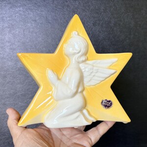 May include: A yellow ceramic star-shaped plaque featuring a white, glossy, relief-sculpted angel kneeling with hands pressed together in prayer. A small, dark purple sticker with the text Royal Copley is affixed to the lower right side.