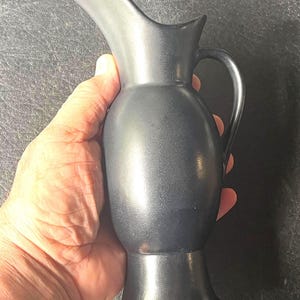 May include: A matte black ceramic pitcher with a smooth, rounded body, a flared base, and a curved handle. The pitcher features a prominent, elongated pouring spout that extends upward from the rim, creating a sleek and continuous silhouette.