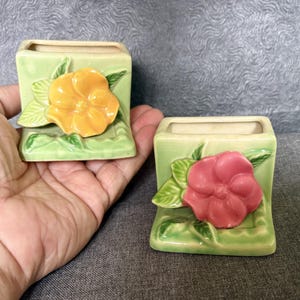 May include: Two square ceramic planters with a light green glazed finish. Each planter features a raised, three-dimensional flower design on the front. One flower is bright yellow with green leaves, and the other is pink with green leaves.
