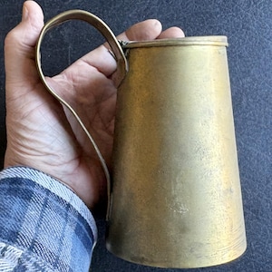 May include: A vintage brass pitcher with a curved handle. The pitcher has a cylindrical body that tapers slightly towards the base. The brass has a warm, aged patina. The handle is attached near the top and bottom of the pitcher.