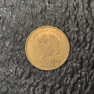 May include: A round, gold-colored coin with the text "GRIZZLY JACK'S GRAND BEAR LODGE" around the edge. The coin features a bear illustration in the center. The coin is set against a textured black surface.