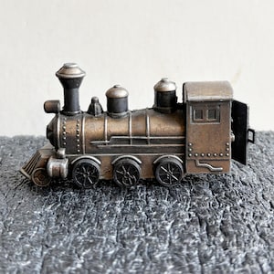 May include: A detailed miniature train engine in shades of bronze and black. The model features a classic steam engine design with a smokestack, multiple wheels, and a small cabin. The train is set against a textured gray surface.