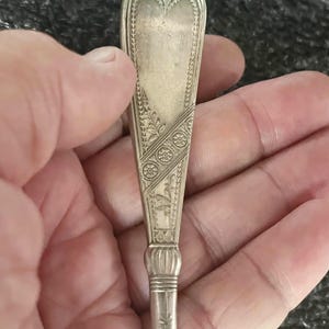 May include: A silver spoon with an ornate design. The spoon's bowl has a heart-shaped top and is decorated with floral and geometric patterns. The handle is slender and features a decorative bulbous section near the base.