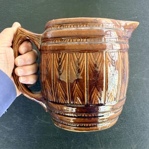 May include: A brown ceramic pitcher with a handle and spout. The pitcher has a textured, barrel-like design with vertical and horizontal bands. The handle has a wood-grain appearance. The pitcher is a warm brown color.