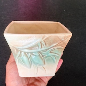 May include: A square ceramic planter with a tapered base, featuring a cream-colored glaze and a raised, embossed leaf pattern in a soft mint green hue. The vessel has a wide, open top and a smooth, matte finish across its exterior surface.