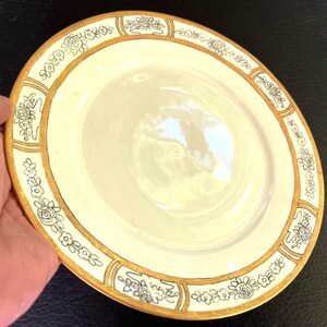 May include: A round ceramic plate with a cream-colored center and a wide border featuring black line-drawn floral patterns. The rim and the sections separating the floral motifs are accented with a metallic gold finish, creating a decorative frame.