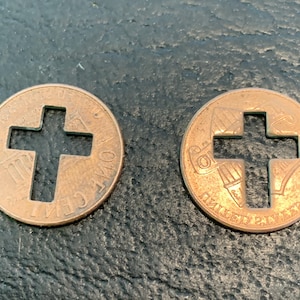 May include: Two copper US one-cent coins with a cross cut out of the center. The coins are on a dark gray surface.