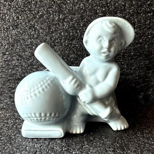 May include: A light blue ceramic figurine of a child holding a baseball bat, leaning against a baseball. The figurine is wearing a hat. The baseball has visible stitching details. The figurine is on a rectangular base.