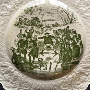 May include: A cream-colored plate with a green transferware design depicting a winter scene with people ice skating. The plate has a decorative, embossed border. The scene includes figures in period clothing, trees, and buildings.