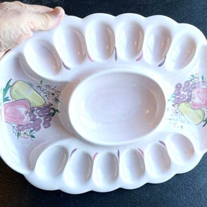 May include: A light pink ceramic serving platter with a scalloped edge and a central oval bowl. The platter has sections for serving and is decorated with fruit illustrations in shades of green, purple, and pink.