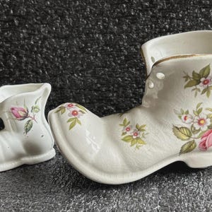 May include: Two white ceramic boot-shaped vases. The larger boot has a gold rim and is decorated with pink roses and floral accents. The smaller boot has a pink rose and green leaves. Both are on a textured surface.