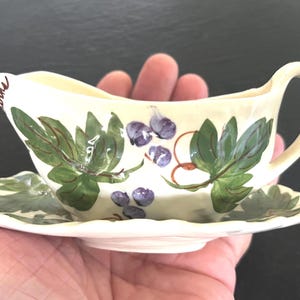 May include: A small, cream-colored gravy boat with a matching saucer. The boat and saucer are decorated with green leaves and purple grapes. The gravy boat has a handle and a spout. A charming addition to any table setting.