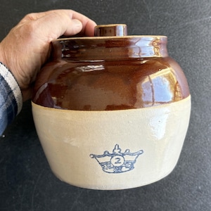 May include: A vintage stoneware crock with a brown glazed top and a cream-colored base. The crock features a blue emblem with the number "2" and a lid. The antique kitchenware is held by a person.