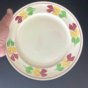 May include: A round ceramic dinner plate with a cream-colored base and a smooth surface. The rim features a repeating pattern of stylized leaf motifs in shades of yellow, green, and maroon, accented by a thin red circular border line.