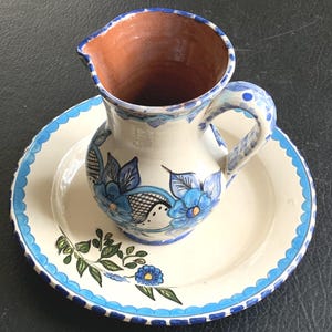 May include: A ceramic pitcher and matching plate set featuring a cream-colored base with hand-painted blue floral patterns, green leaves, and a scalloped blue rim. The pitcher has a terracotta-colored interior and a curved handle for pouring.