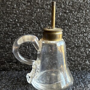 May include: A clear glass oil can with a handle and a brass top. The can has a spout and a small opening for filling. The handle is curved and attached to the side of the can. The oil can is likely vintage.