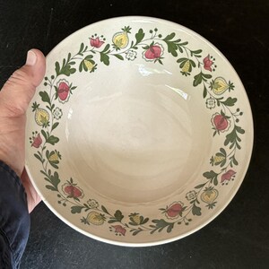 May include: Cream-colored ceramic bowl with a floral design. The rim features a garland of red, yellow, and white flowers with green leaves. The bowl is held by a person's hand, showcasing its size and shape. Ideal for serving soups or salads.