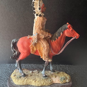 May include: A detailed figurine of a Native American chief riding a red horse. The chief is wearing a feathered headdress and traditional clothing. The horse has a black mane and tail. The figurine is mounted on a wooden base with a textured ground.