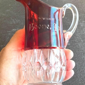 May include: A small glass pitcher featuring a deep red upper section and a clear, diamond-cut patterned lower base. The red portion is etched with the text Souvenir Boone, Ia. It includes a clear glass handle and a curved pouring spout.