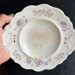 May include: A white ceramic serving dish with a scalloped edge and gold accents. The dish has a floral design with purple and gold flowers. It has two handles on the sides.