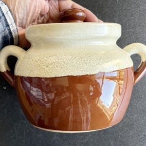 May include: A two-handled, lidded ceramic pot with a speckled beige top and a glossy brown base. The pot has a rounded shape with two curved handles and a small, round knob on the lid. The pot is likely vintage.