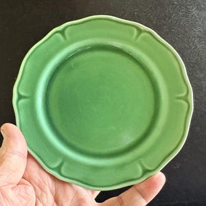 May include: A round, green ceramic plate featuring a scalloped edge and a subtle, fine crackle glaze finish across the surface. The plate has a smooth, flat center and a raised, decorative rim with distinct indentations.